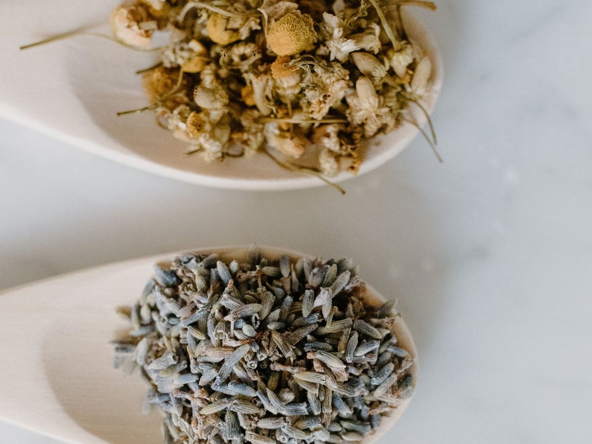 Colheres de madeira com camomila seca, botões de lavanda e folhas secas sobre mármore branco